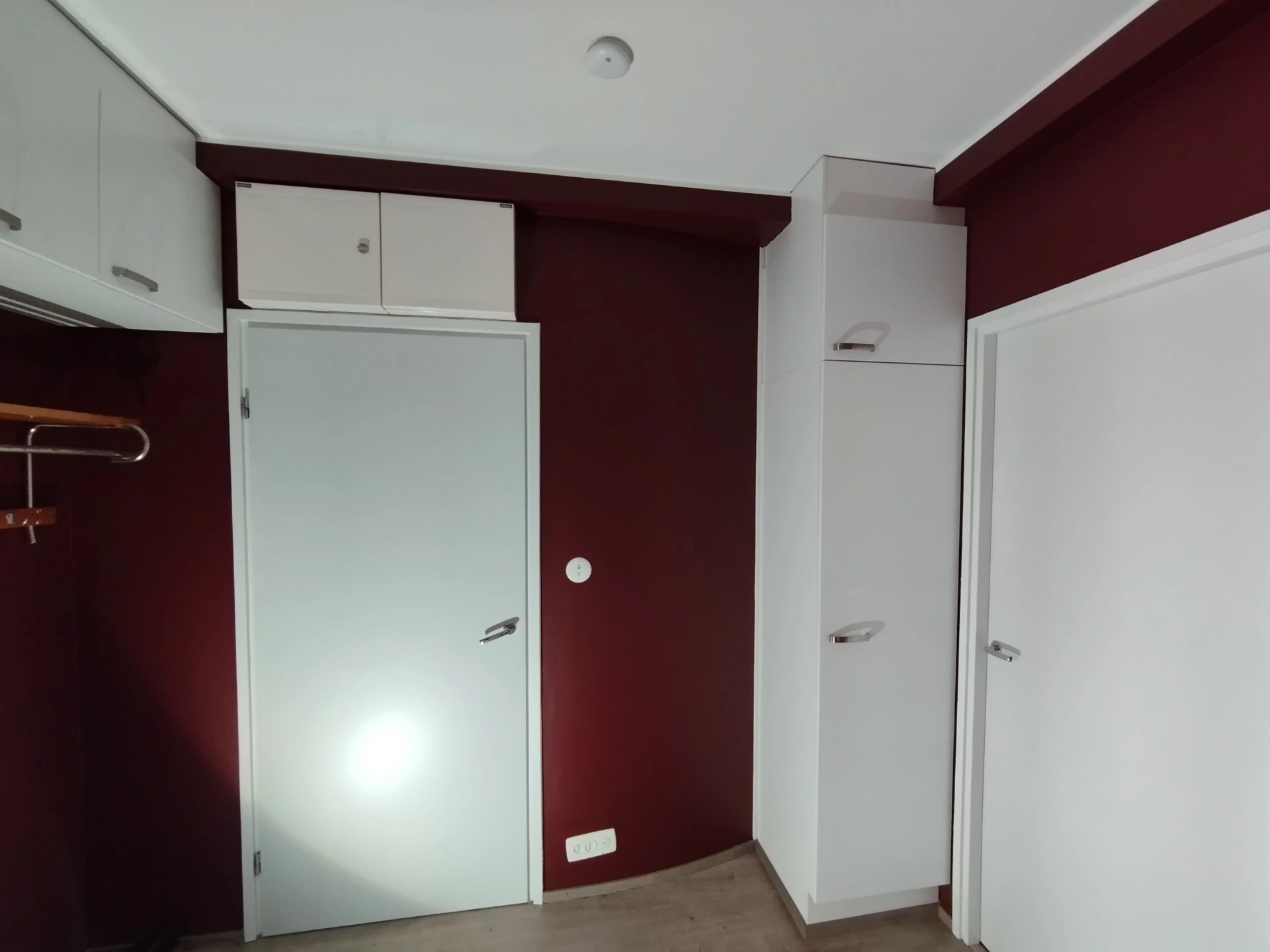Huoneessa on valkoiset kaapit, ovi ja tummanpunainen seinä. Katossa on valkoinen valaisin.