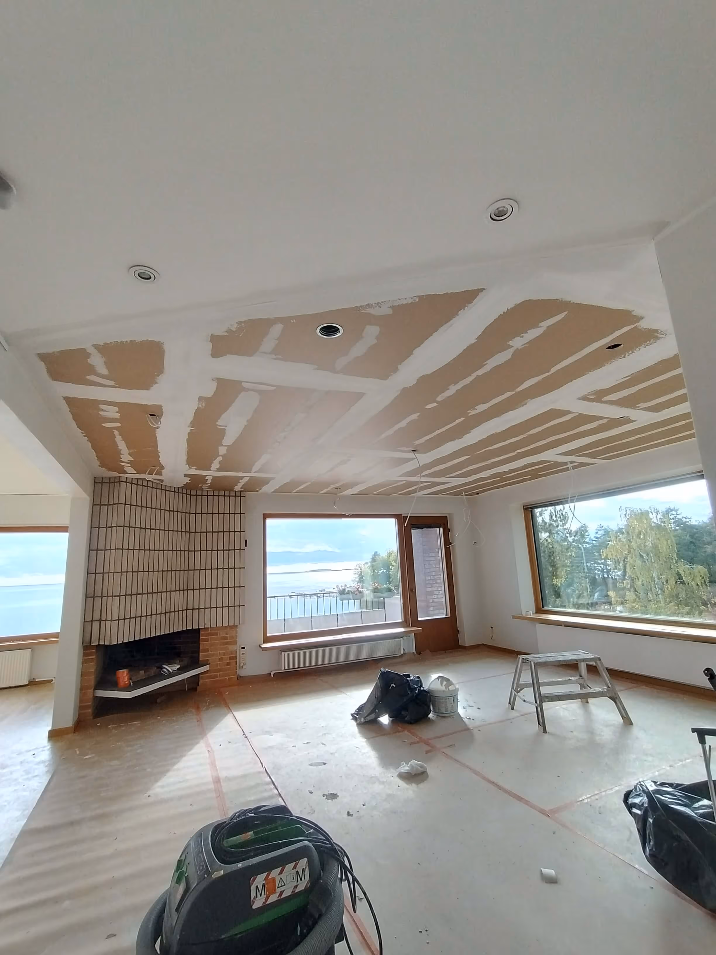 Huoneessa on keskeneräinen katto, jossa on näkyvissä kipsilevy ja saumaus, sekä suuri tiilitakka ja näkymät merelle ja luontoon suurista ikkunoista.