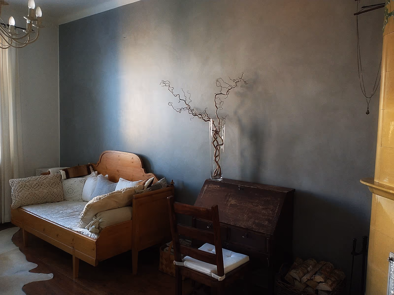 Huoneessa on puinen sohva, kirjoituspöytä, tuoli ja maljakko oksilla harmaata seinää vasten.