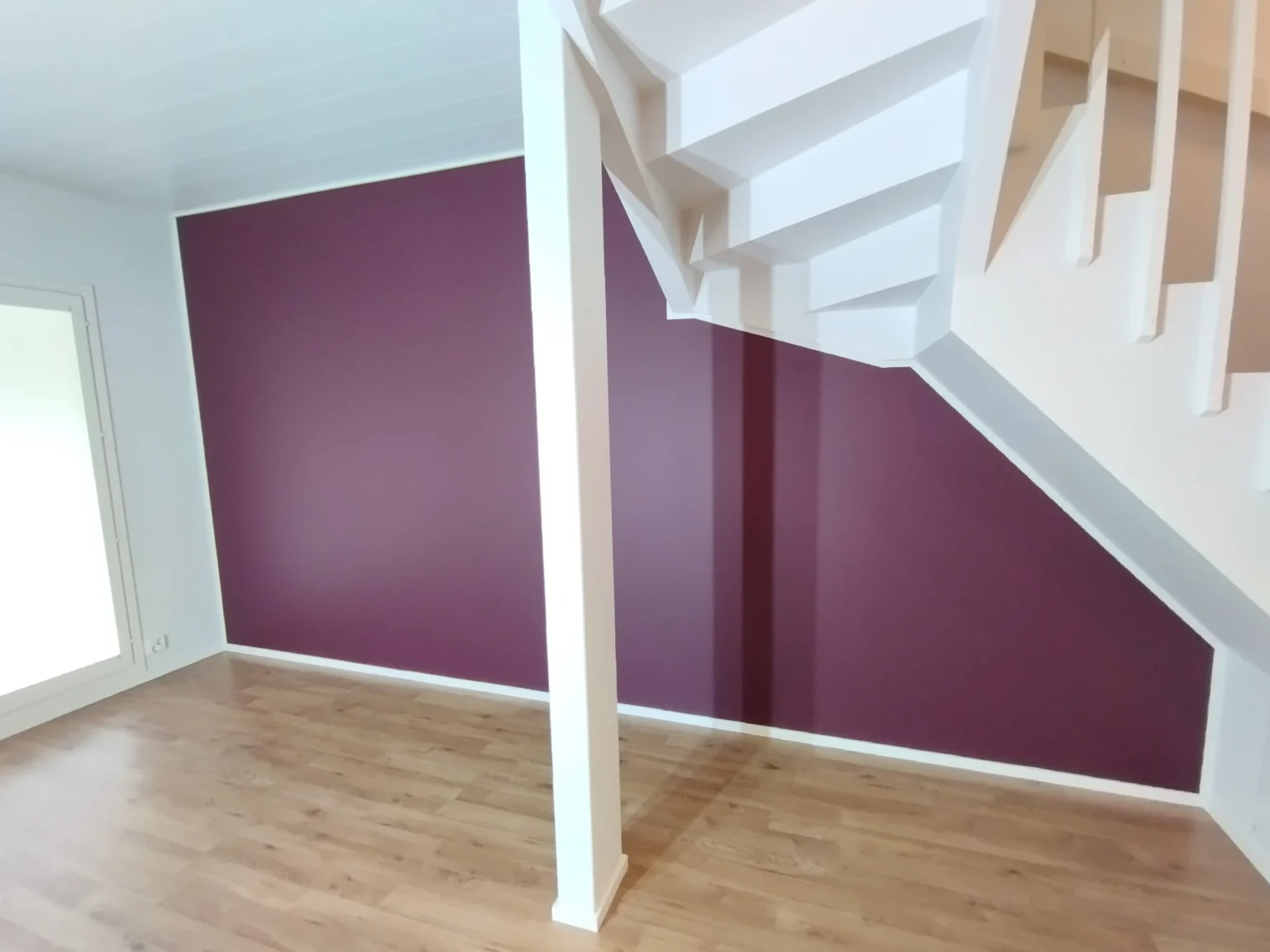 Sisätila, jossa on puulattia, viininpunainen seinä ja valkoiset portaat. Huone on valoisa ja avara.