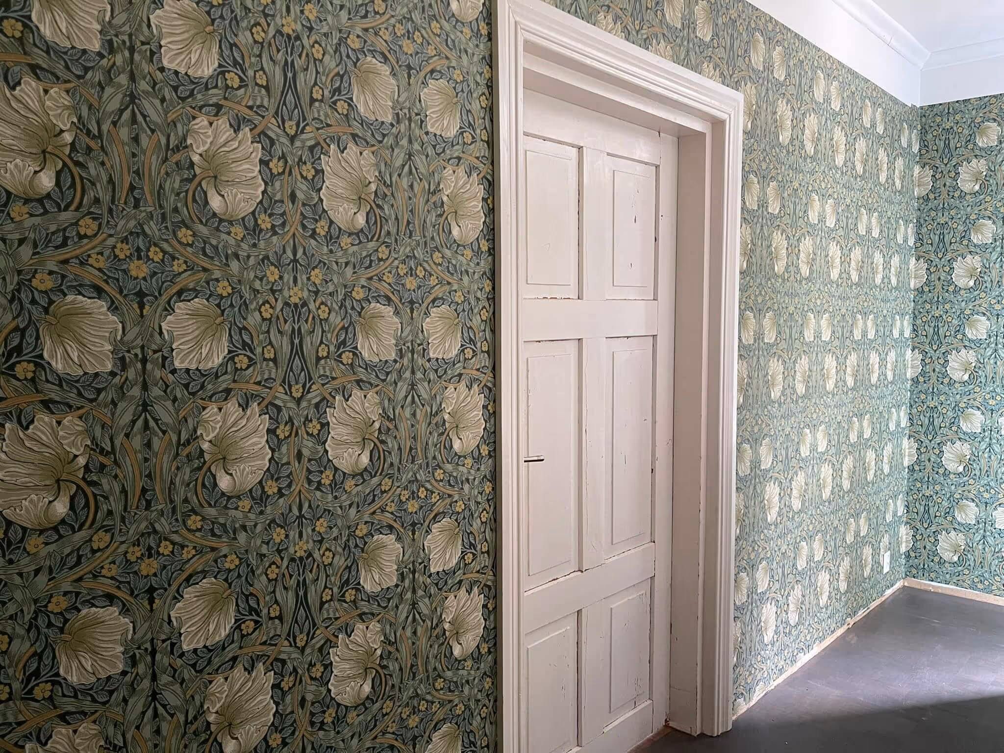 Huoneessa on kukkatapetti, valkoinen ovi ja tumma lattia. Tapetti on vihreä ja siinä on suuria, vaaleita kukkia.