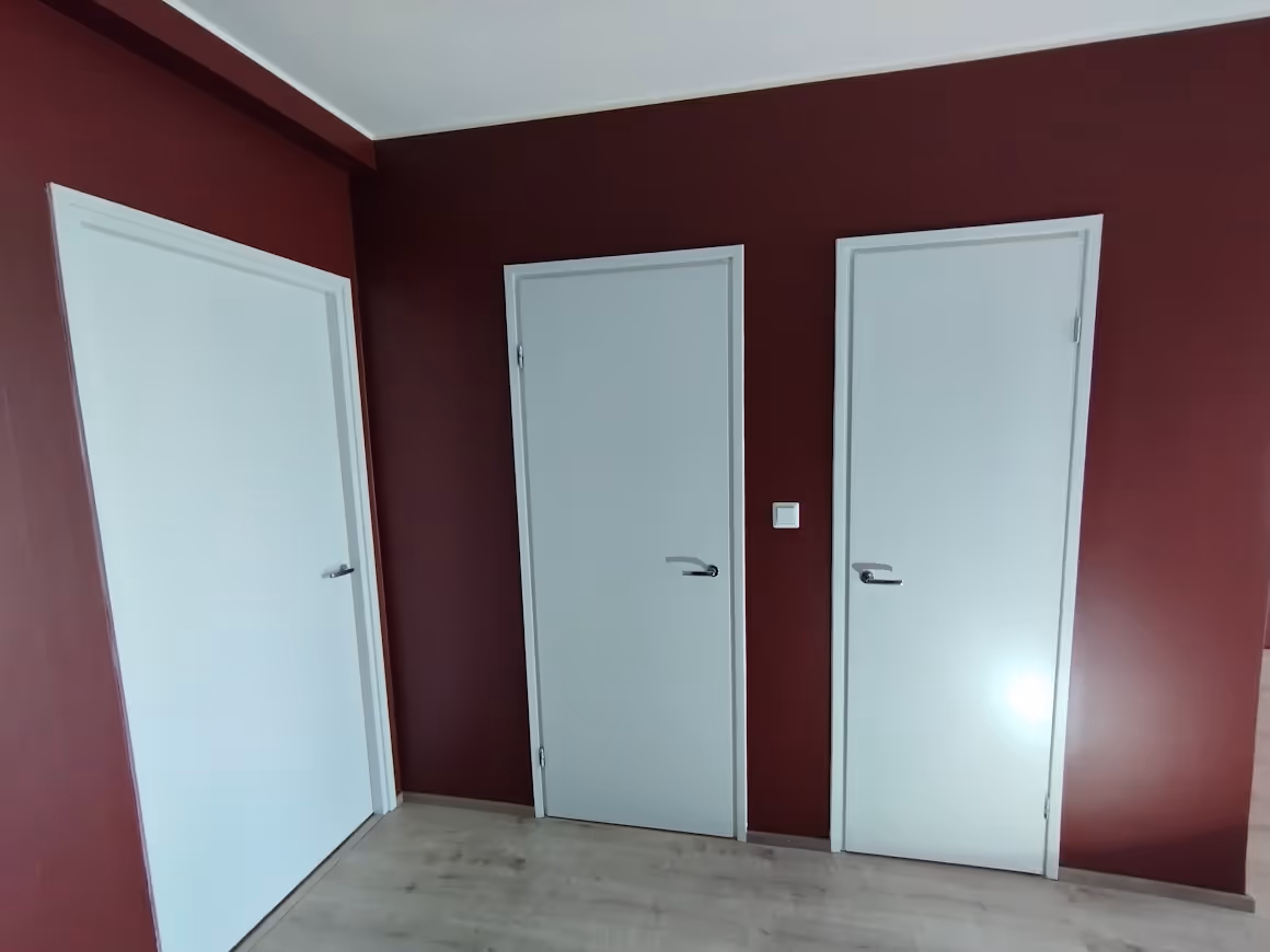 Kolme valkoista ovea viininpunaisella seinällä valoisassa huoneessa, jossa on vaalea lattia.