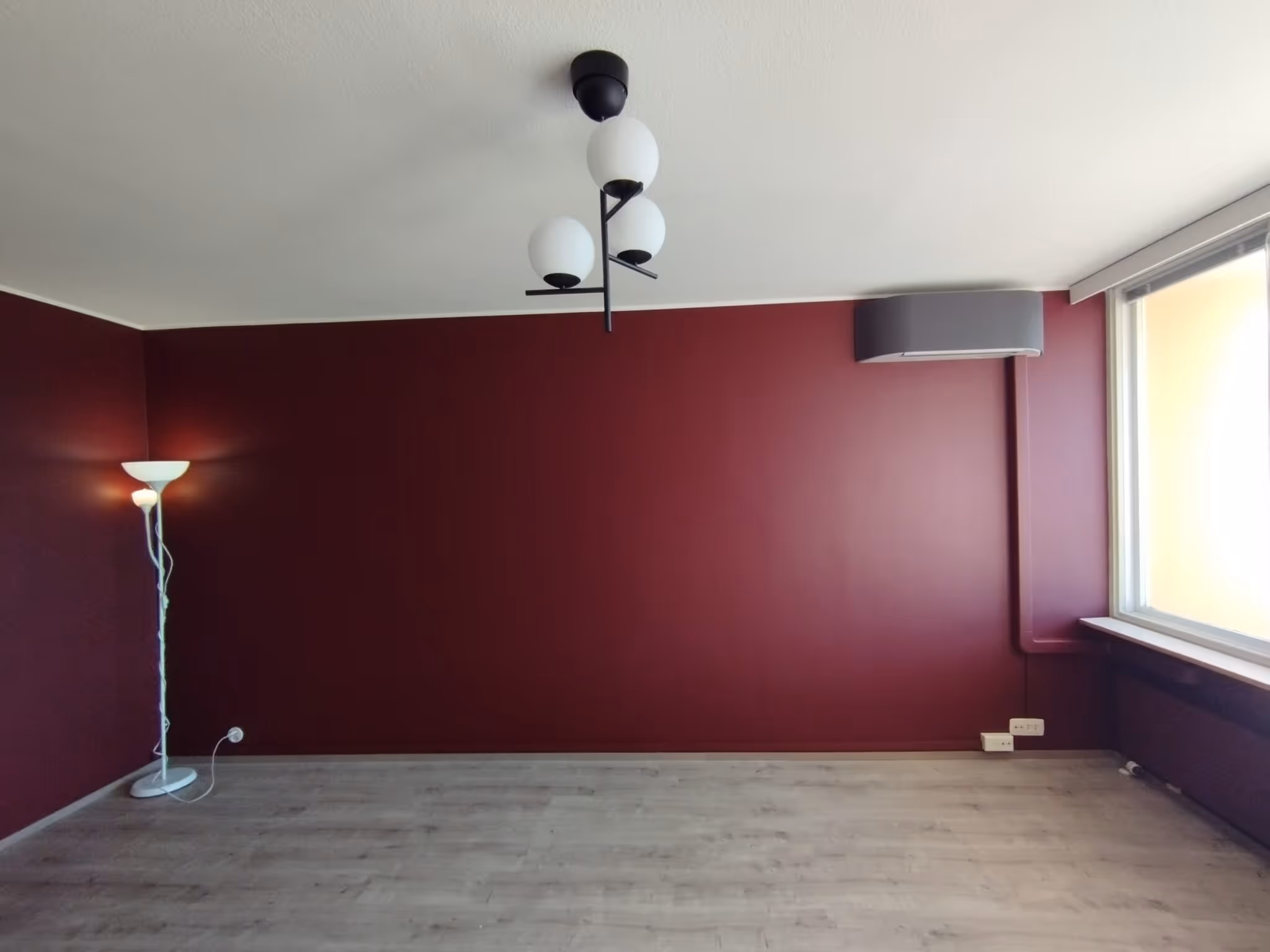 Huoneessa on punaiset seinät, vaalea puulattia, valkoinen katto, moderni kattovalaisin ja lattiavalaisin. Ikkunasta tulee valoa.