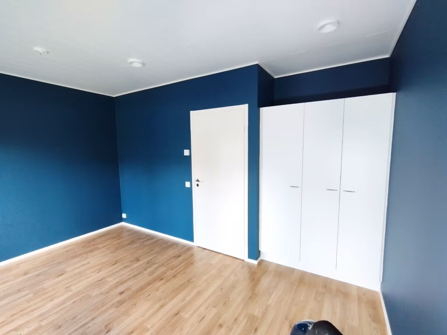 Huoneessa on siniset seinät, vaalea puulattia, valkoinen ovi ja kolmeovinen vaatekaappi. Huone on valoisa.