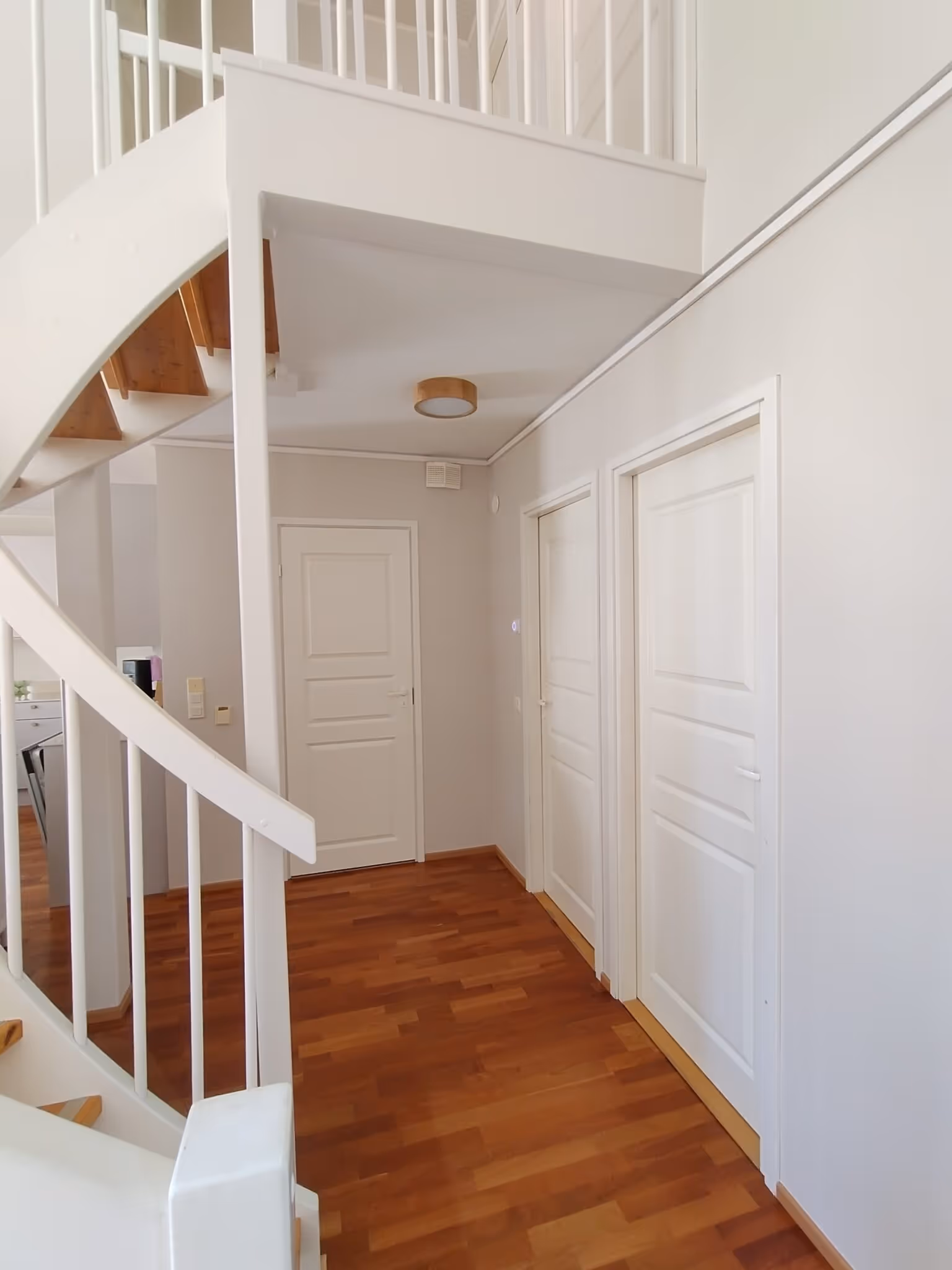 Sisätiloissa on valkoiset portaat, puulattia ja valkoiset ovet. Huoneessa on myös valaisin katossa.