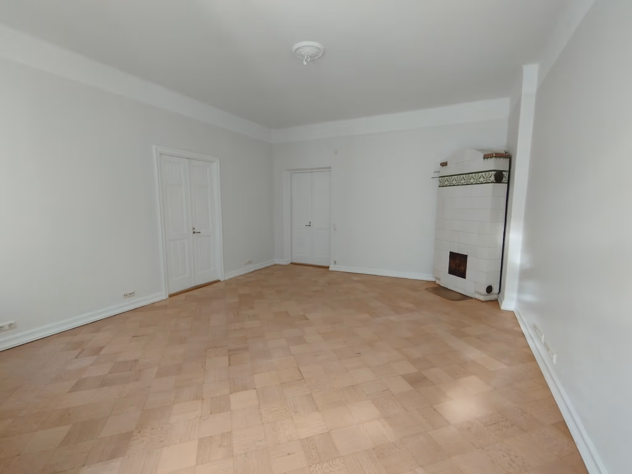 Tyhjä huone, jossa on puulattia, valkoiset seinät, kakluuni ja kaksi valkoista ovea. Huone on valoisa.