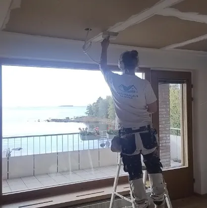 Sisätiloissa tikkailla seisova henkilö asentaa valaisinta kattoon, jossa on ruutukuviointi. Ikkunasta näkyy merimaisema.