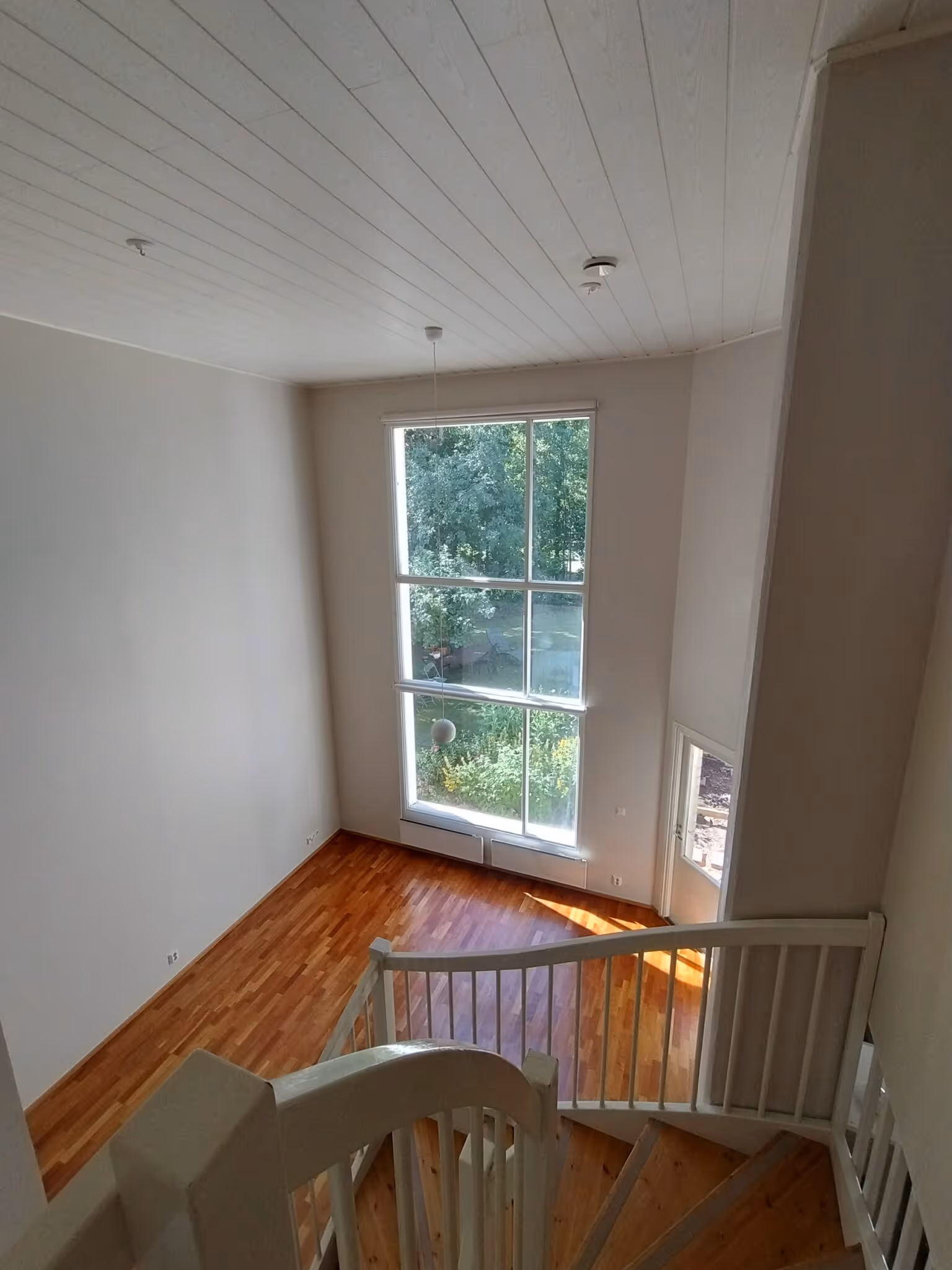 Valoisa sisätila, jossa on puulattia, suuri ikkuna, valkoiset seinät ja katto sekä puuportaat valkoisella kaiteella.