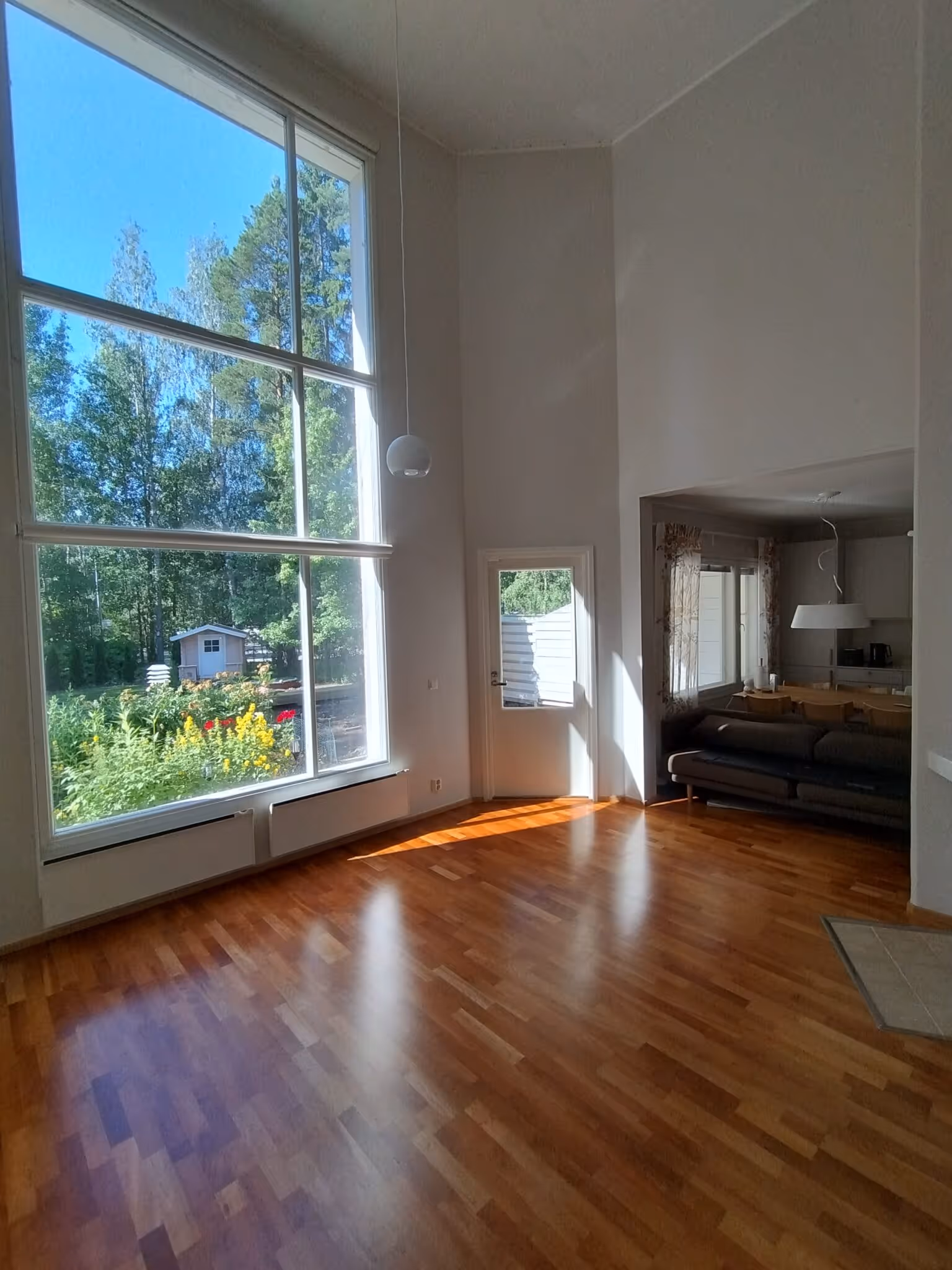 Valoisa olohuone, jossa suuri ikkuna, puulattia, sohva ja näkymä puutarhaan.
